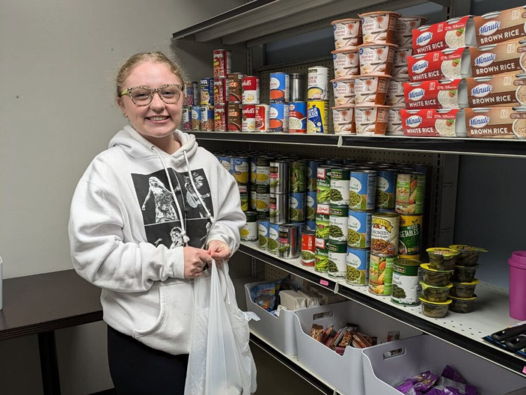 Student smiles next to food items on a pantry shelf.