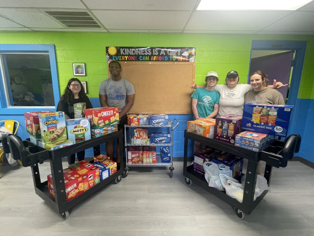 A group of volunteers pose proudly behind their service work of donated food items in colorful room.